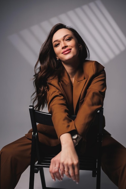 A pretty young woman with a smile on her face and wearing a brown classic suit is sitting on a chair on a gray background Beautiful woman in a jacket and with makeup on her face