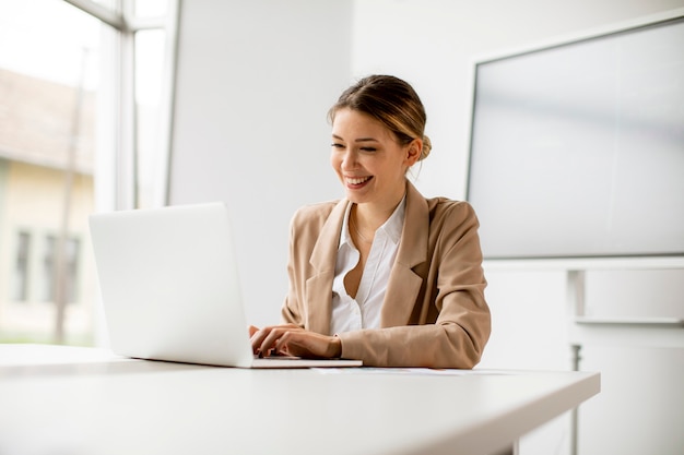 Pretty young woman working on laptop in bright office with big screen behind her