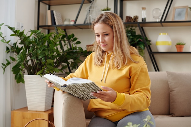 Pretty young woman in yellow hoodie resting on sofa at home and reading interesting book