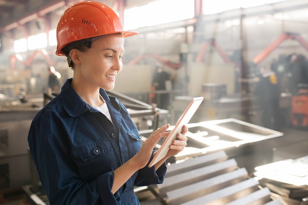 Pretty yung female engineer or technician in uniform and helmet surfing in the net while looking for online data