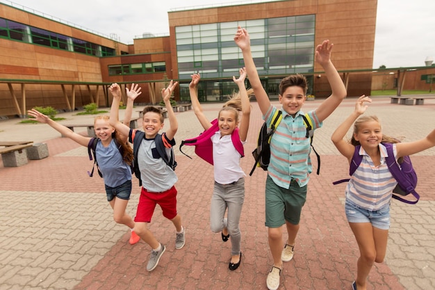 primary education, friendship, childhood and people concept - group of happy elementary school students with backpacks running and waving hands outdoors