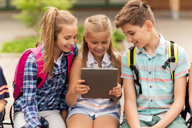 primary education, friendship, childhood, technology and people concept - group of happy elementary school students with backpacks sitting on bench and talking outdoors