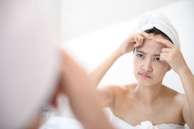 Problem skin Concerned young asian women popping pimple on cheek while standing near mirror in bathroom young asian women with acne