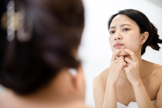 Problem skin Concerned young asian women popping pimple on cheek while standing near mirror in bathroom young asian women with acne