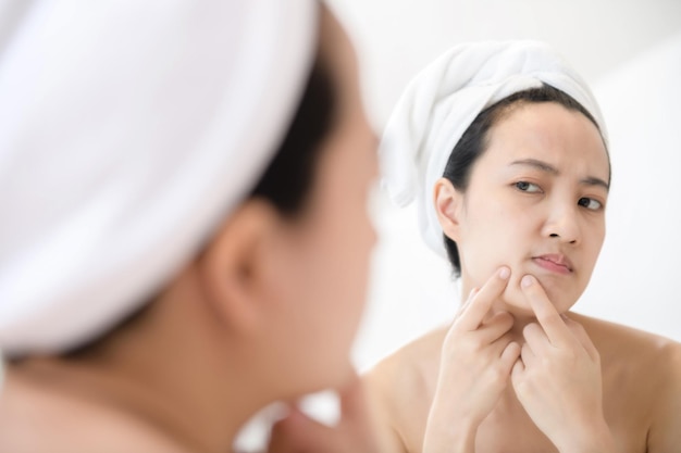 Problem skin Concerned young asian women popping pimple on cheek while standing near mirror in bathroom young asian women with acne