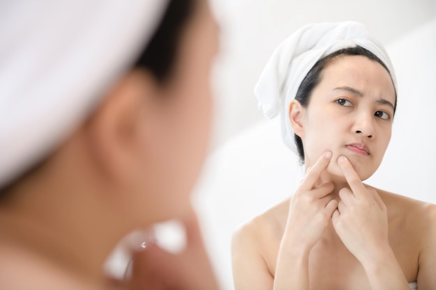 Problem skin Concerned young asian women popping pimple on cheek while standing near mirror in bathroom young asian women with acne