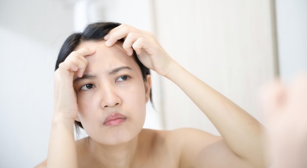 Problem skin Concerned young asian women popping pimple on cheek while standing near mirror in bathroom young asian women with acne