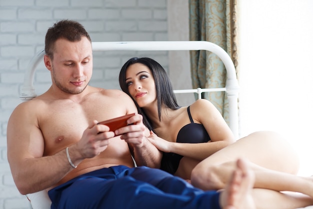 Problems in the family. Angry young woman lying on the bed, against her husband, who plays the game on the phone.