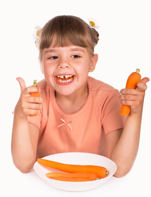 Problems of pediatric dentistry Little girl in an orange tshirt and flowers in her hair eats a carrot on a white background