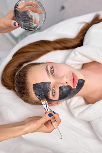 Procedure for applying black mask on fresh skin of face of beautiful woman, top view. Spa treatments, skin care, beauty concept. Attractive female look at camera, close-up photo. copy space