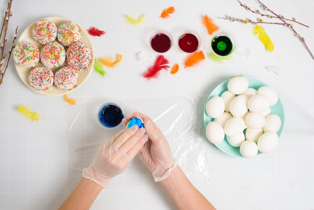 process of coloring eggs for Easter in blue and red colors. a woman in gloves paints eggs with dye from a jar, top view, willow branches and colorful feathers decor.