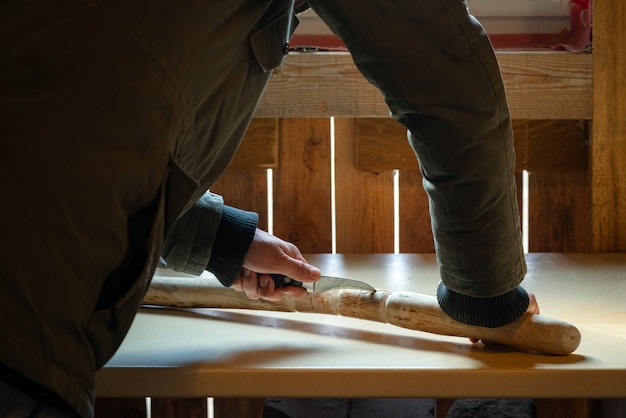 Process of man making wooden walking stick indoors during quarantine. Carving wood stick on the table using knife