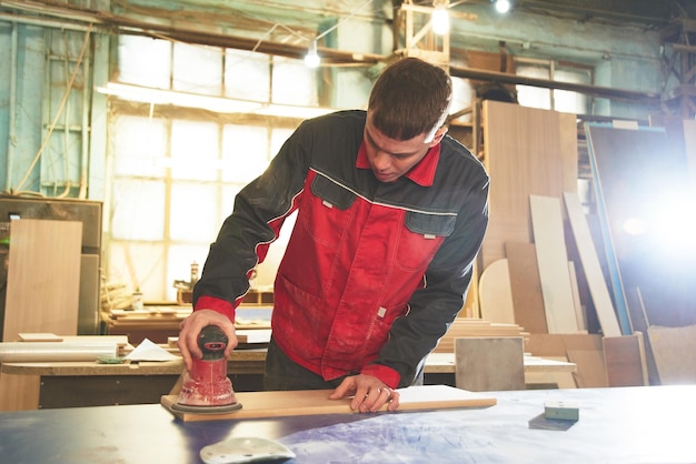 Process of production and manufacture of wooden furniture in furniture factory Worker carpenter man in overalls processes wood on special equipment