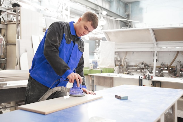 Process of production and manufacture of wooden furniture in furniture factory Worker carpenter man in overalls processes wood on special equipment