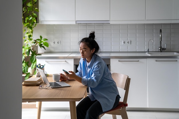 Procrastinating Asian woman browsing social media feed on phone sits at kitchen table with laptop