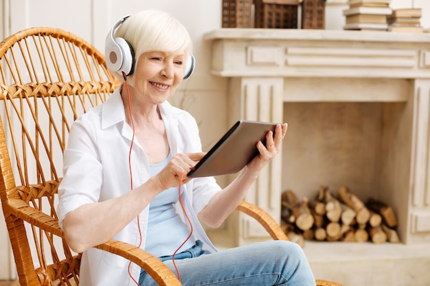 Productive intelligent elderly woman using her tablet for enjoying some nice videos while sitting in a chair and wearing headphones