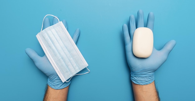 Products to stay safe during pandemic covid19 quarantine. Hands in blue gloves holding sanitiser gel, soap, medical face masks on blue abstract background. Protection against coronavirus, banner size