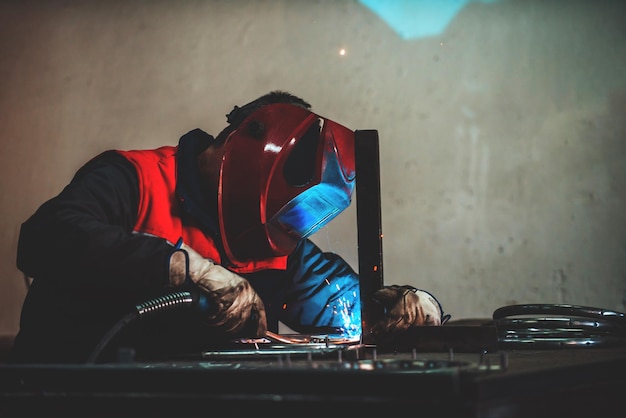 Profesional welder in protective uniform and mask welding metal pipe on the industrial table with other workers behind in the industrial workshop