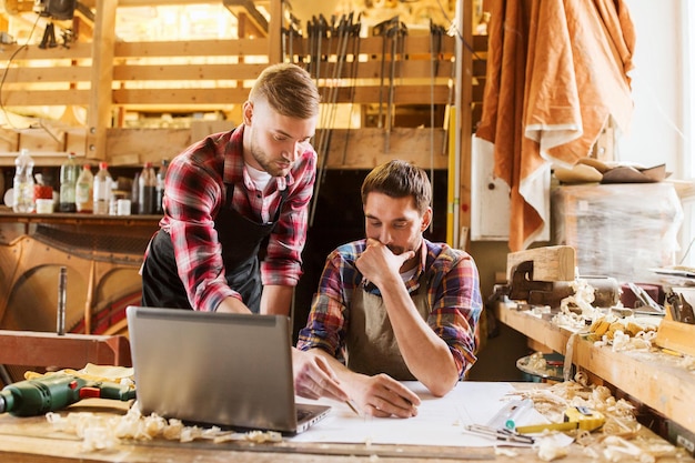 profession, carpentry, technology and people concept - two carpenters with laptop computer and blueprint at workshop