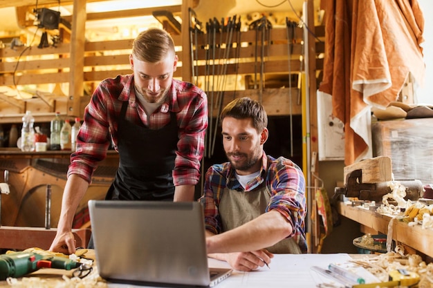 profession, carpentry, technology and people concept - two carpenters with laptop computer and blueprint at workshop