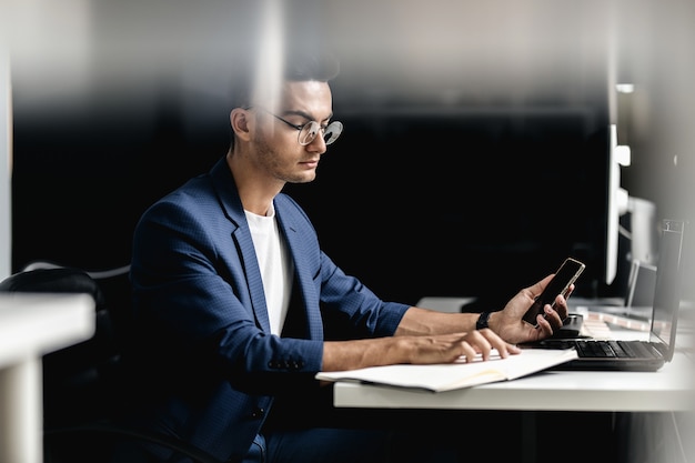 Professional architect in glasses dressed in blue checkered jacket sits in the armchair near the desk with laptop and uses the phone .