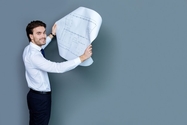 Photo professional architect. joyful nice handsome man smiling and looking at you while holding a scheme