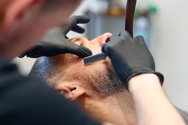 Professional barber shaves customer beard with straight razor.