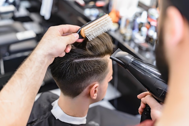 Professional barber styling hair of his client. Barber makes hair styling with a hair dryer.