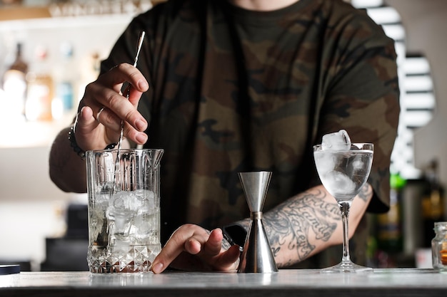 Professional bartender stirs cold cocktail with spoon Several bottles of alcoholic drinks and glasses standing on the bar counter