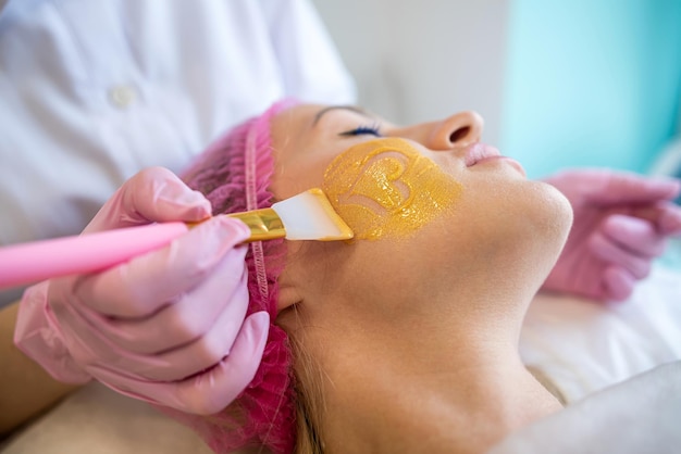 A professional beautician applies a golden healing mask to a woman's face Cosmetology and facial skin care in a beauty salon Cosmetic procedure