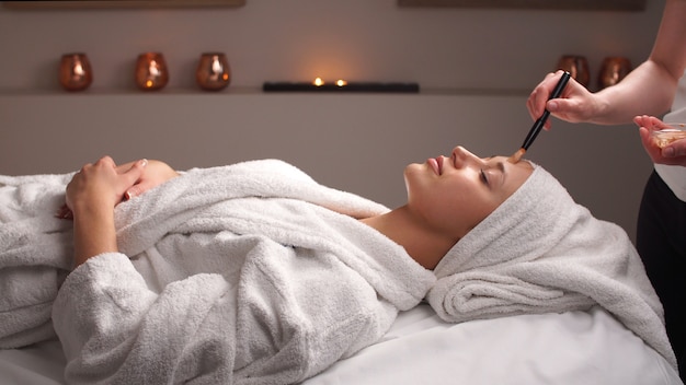 Professional beautician applying cosmetic mask on female face at beauty salon. Woman is having cosmetic treatment at spa salon.