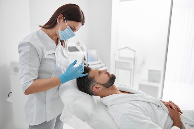 Professional beautician holds syringe with filler above forehead of client in cosmetology clinic