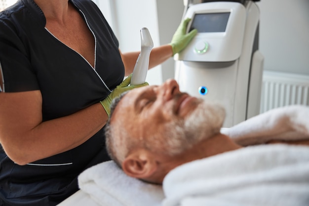 Photo professional beautician starting a machine for photofacial treatment while providing skincare for an elderly man