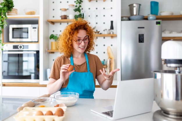 Professional beautiful happy young woman is blogging for her kitchen channel about healthy living in the kitchen of her home and looking on camera on a laptop