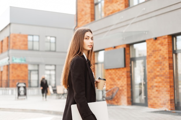 Professional business manager woman with black elegant clothes with laptop and cup of coffee walks in the city