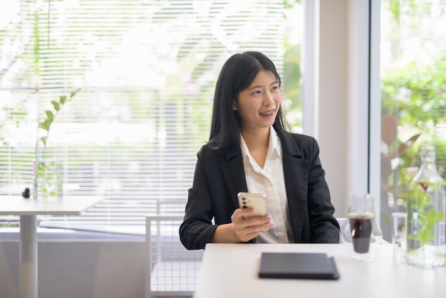 Professional businesswoman working on her smartphone in a bright modern cafe with coffee and greenery looking through window while smiling