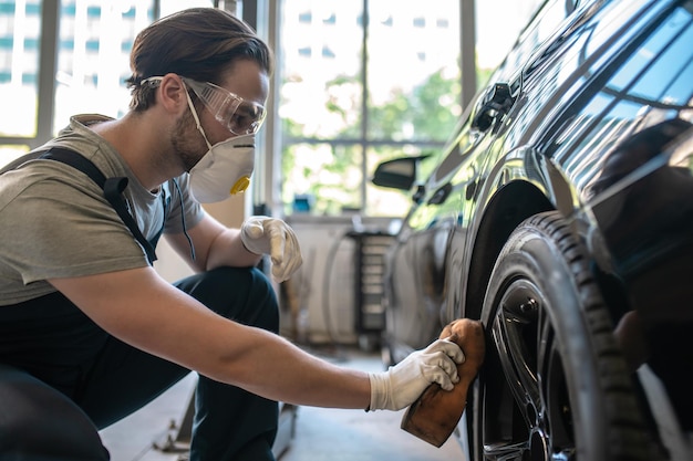 Professional car detailer in the automotive respirator buffing the automobile wheel surface with a foam sponge