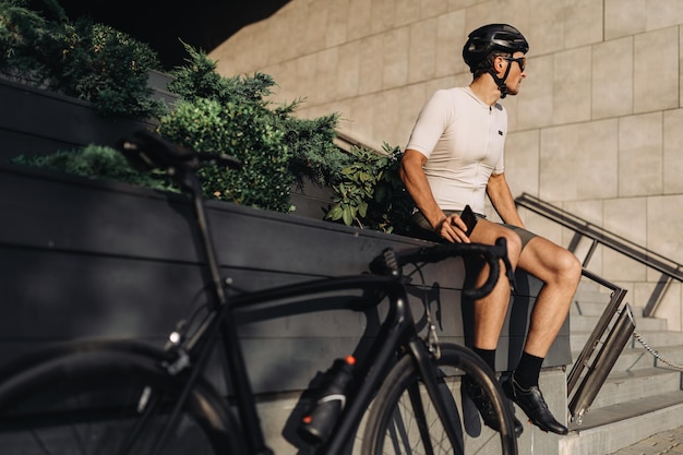 Professional caucasian cyclist with smartphone in hands looking aside while sitting near his bike on street Training outdoors during summer time Technology for lifestyles