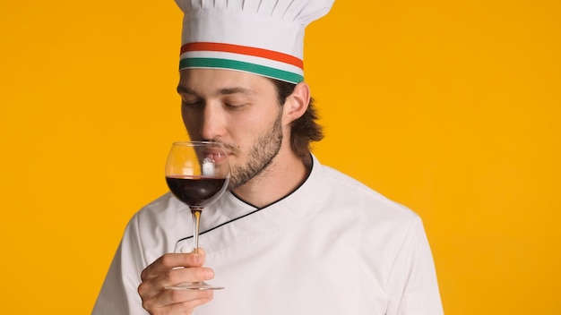 Professional chef dressed in uniform holding glass of wine isolated on yellow background Man sommelier sniffing wine