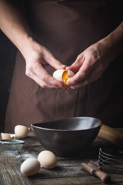 Professional chef hands are breaking an egg into bowl to make dough on wooden table, over dark