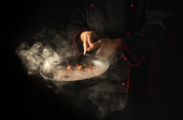 Professional chef prepares food in a frying pan with steam on a black background