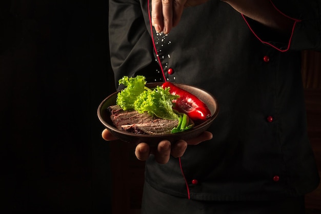 A professional chef sprinkles salt on a sliced beef steak and vegetables in a plate