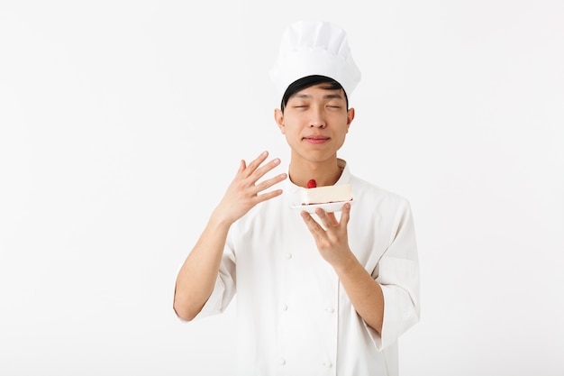 professional chinese chief man in white cook uniform smiling at camera while holding platter with tasty cheesecake isolated over white wall