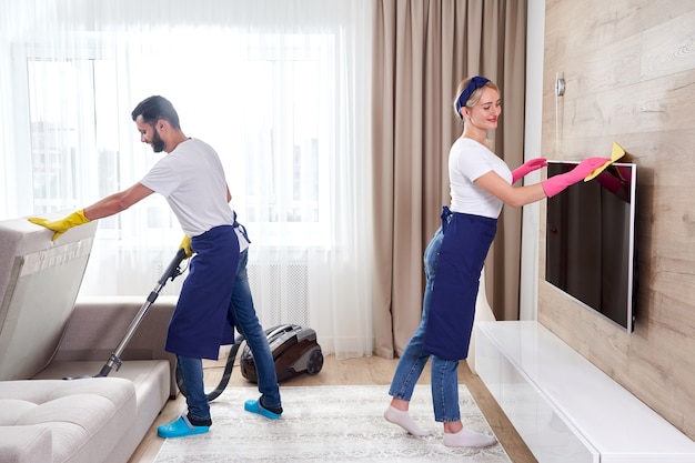 Professional cleaners in blue uniform washing floor and wiping dust from the furniture in the living room of the apartment. Cleaning service concept