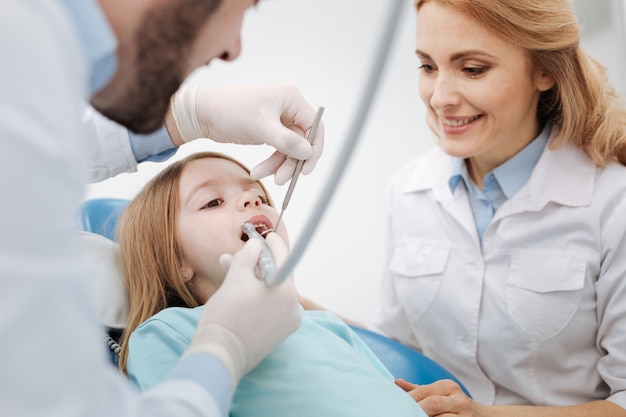 Professional competent medical colleagues examining little patients teeth and running a routine checkup while his colleague comforting her