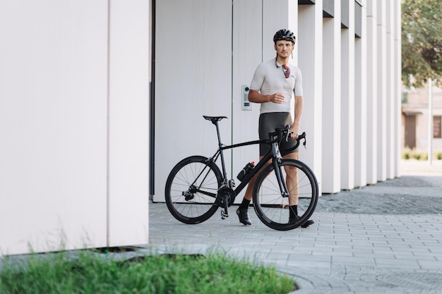 Professional cyclist in sport outfit and glasses posing with his black bike near modern building Caucasian man spending leisure time for active workout outdoors