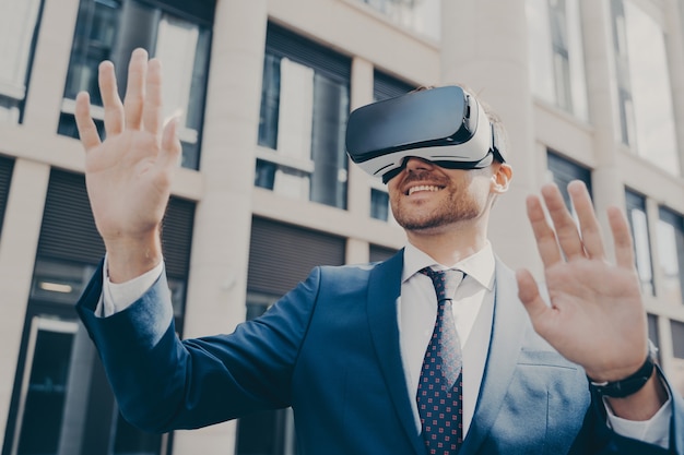 Professional excited office worker in blue suit standing outside alone and using VR glasses to visualize projects, holding something in virtual reality with his hands, building in background