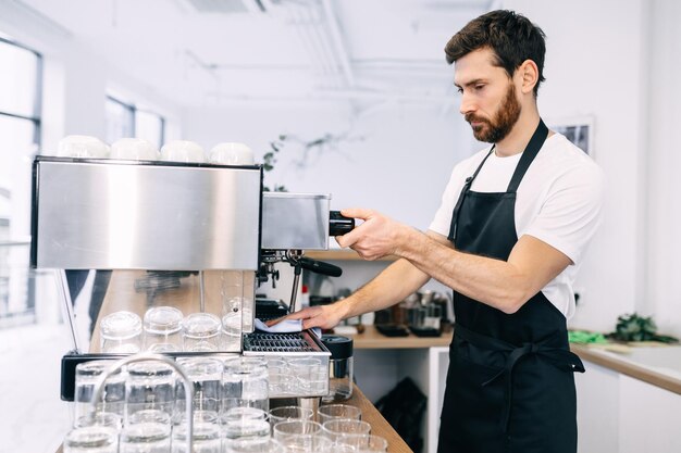 A professional experienced barista prepares coffee in a coffee machine wearing a black apron and white Tshirt