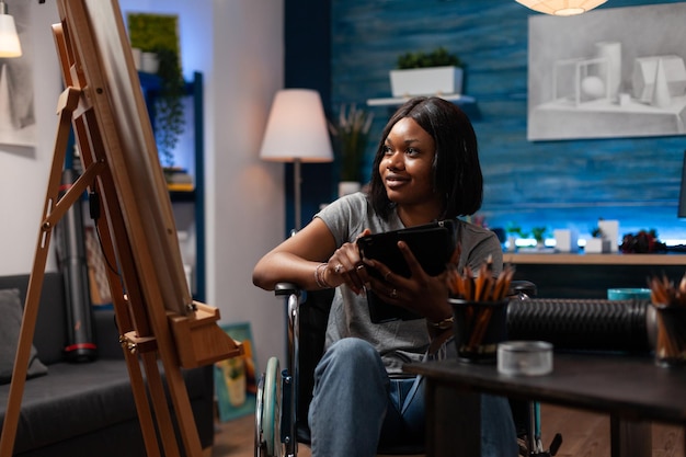 Professional female artist looking for inspiration on internet to create pencil drawing on blank canvas in art studio. African american woman with artistic talent reviewing art notes on tablet.