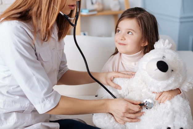 Professional female doctor using stethoscope and examining fluffy bear while sitting on the couch